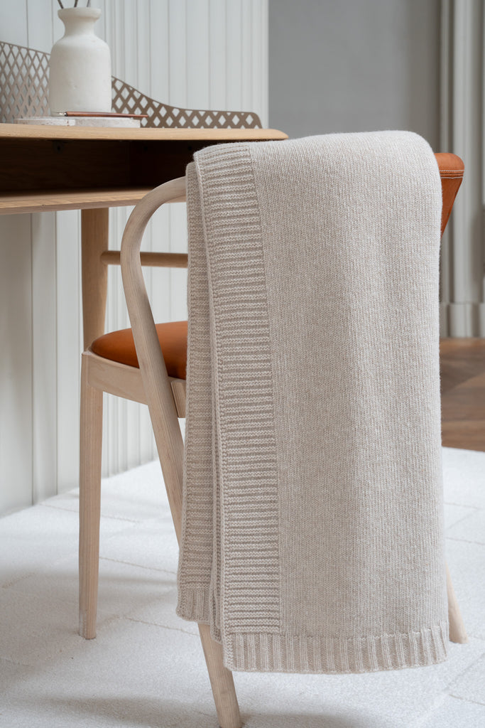 Gladgebreide beige plaid van 100% merinowol, 130 x 170 cm, elegant over een stoel gehangen in Scandinavische interieur met een bureau op de achtergrond.