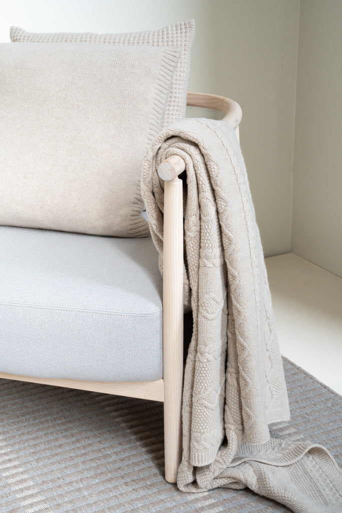 Vlechtmotief beige plaid van 100% merinowol, 130 x 170 cm, nonchalant over de leuning van een houten loungestoel gedrapeerd in een sereen Scandinavisch interieur.