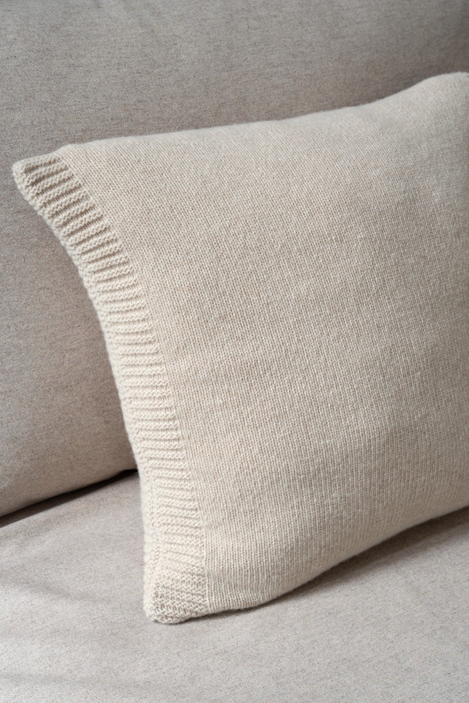 Sfeerbeeld van een beige sierkussen met glad breisel van 100% merinowol, 50 x 50 cm, subtiel geplaatst op een neutrale zetel.
