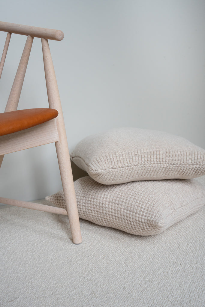 Beige sierkussens van 100% merinowol met wafel- en glad breisel, gestapeld op de vloer in een Scandinavisch interieur naast een houten stoel met leren zitting.