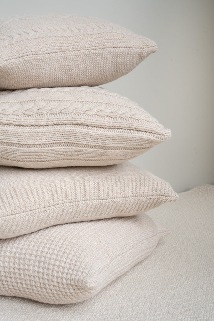 Gestapelde sierkussens van beige merinowol met diverse patronen: wafel, kabel (Aran) en gladbreisel, gefotografeerd op neutrale achtergrond.