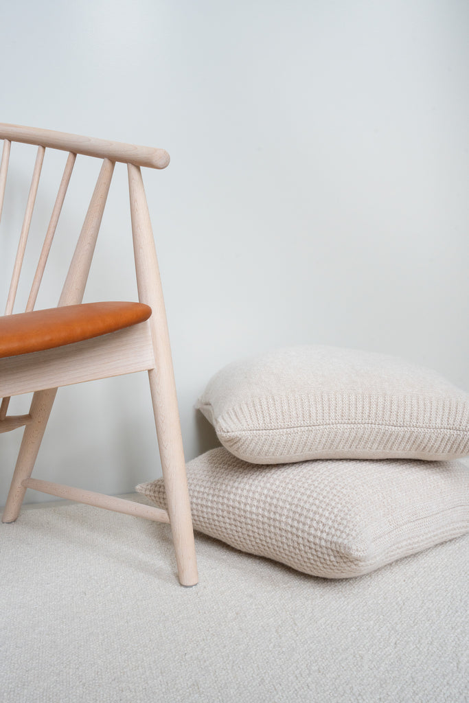 Beige 100% merinowollen sierkussens in Scandinavisch interieur, op de grond naast een houten stoel met leren zitting en neutrale achtergrond.