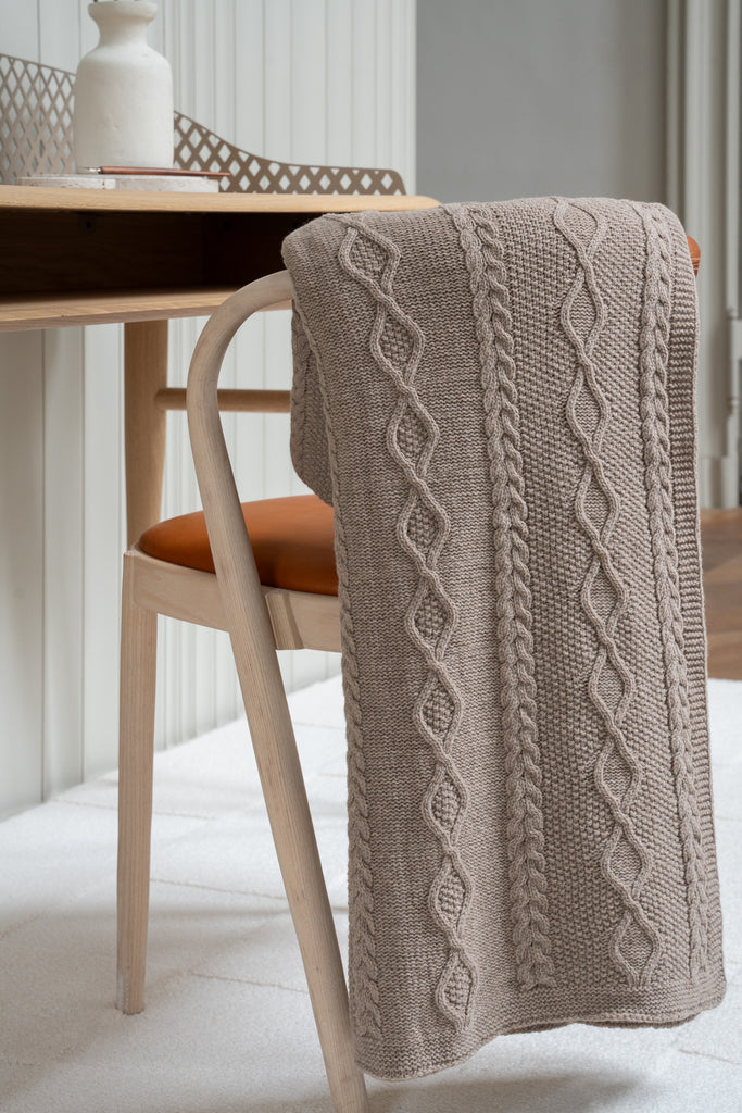 Vlechtmotief bruine plaid van 100% merinowol, 130 x 170 cm, elegant over een stoel gehangen in een Scandinavisch interieur met een bureau op de achtergrond.