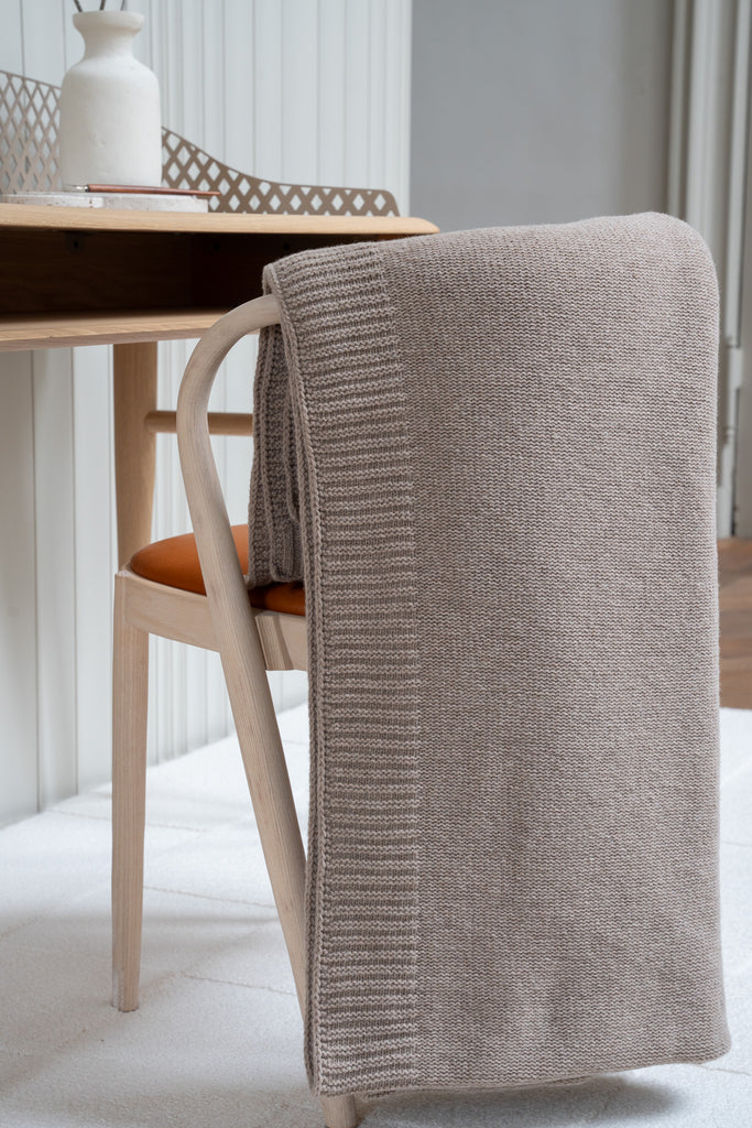Gladgebreide bruine plaid van 100% merinowol, 130 x 170 cm, elegant over een stoel gehangen in Scandinavische interieur met een bureau op de achtergrond.