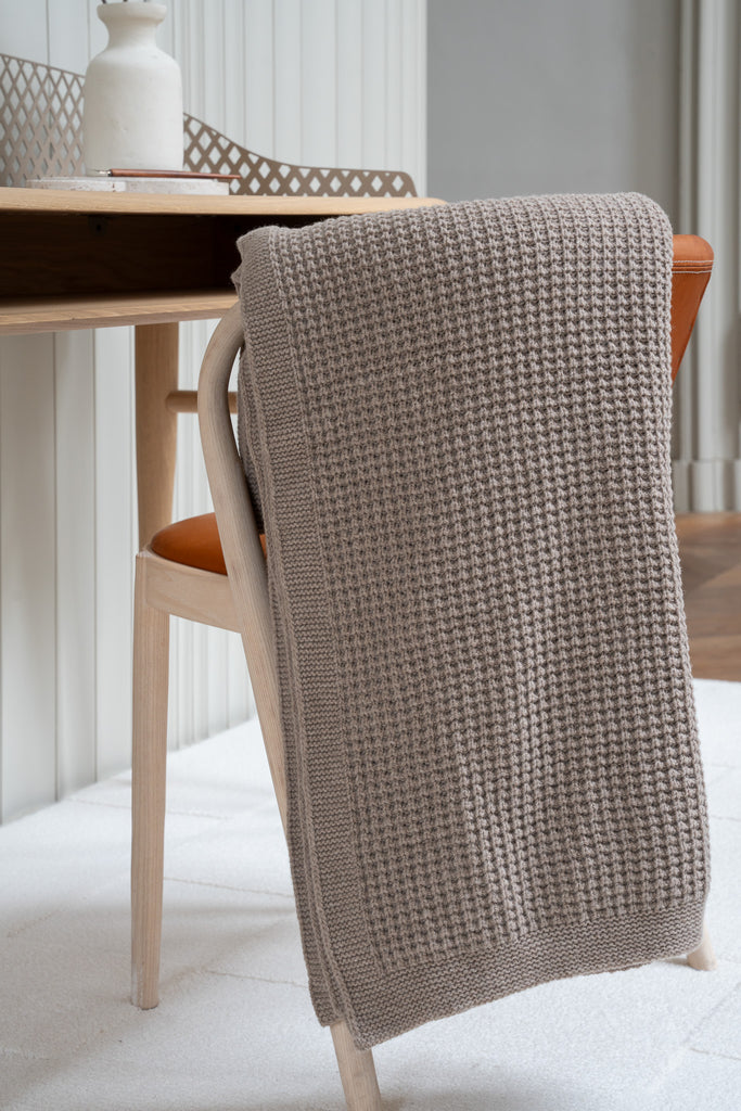 Bruine plaid van 100% merinowol met wafelpatroon, 130 x 170 cm, elegant over een stoel gehangen in Scandinavisch interieur met bureau op de achtergrond.