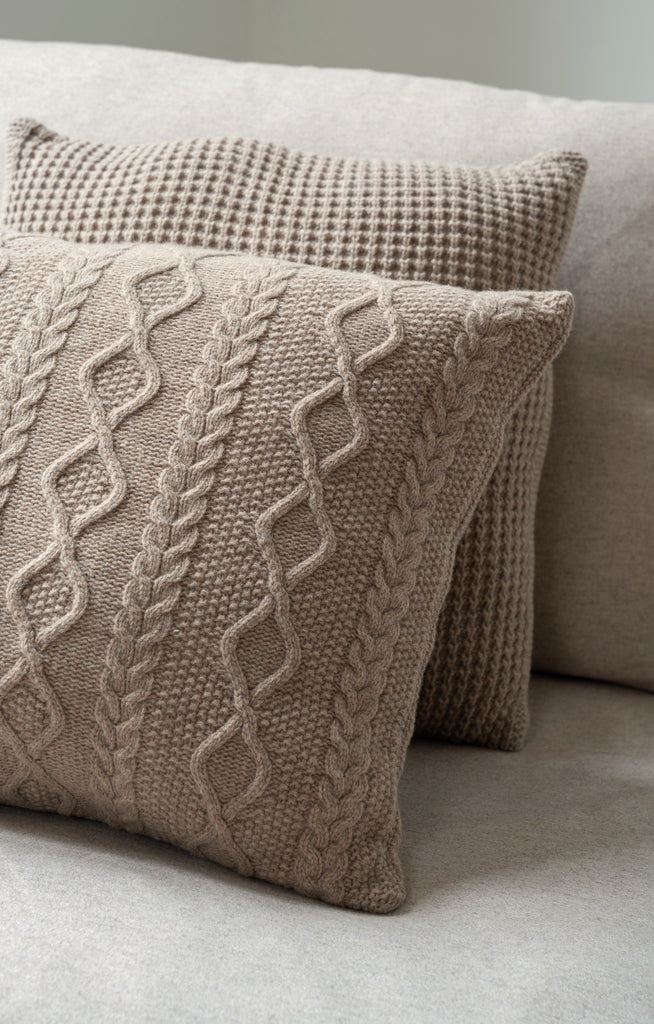 Sfeerbeeld van gestapelde bruine sierkussens van 100% merinowol met wafel- en kabelpatronen (Aran), subtiel gestyled op een stijlvolle zetel.