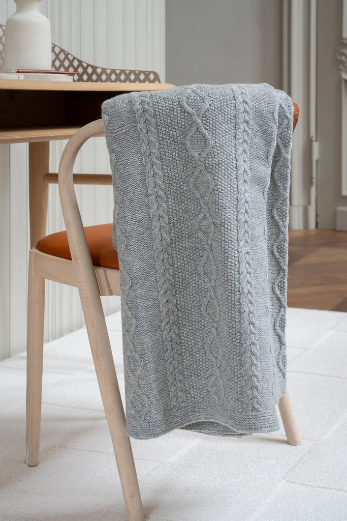 Vlechtmotief grijze plaid van 100% merinowol, 130 x 170 cm, elegant over een stoel gehangen in een Scandinavisch interieur met een bureau op de achtergrond.