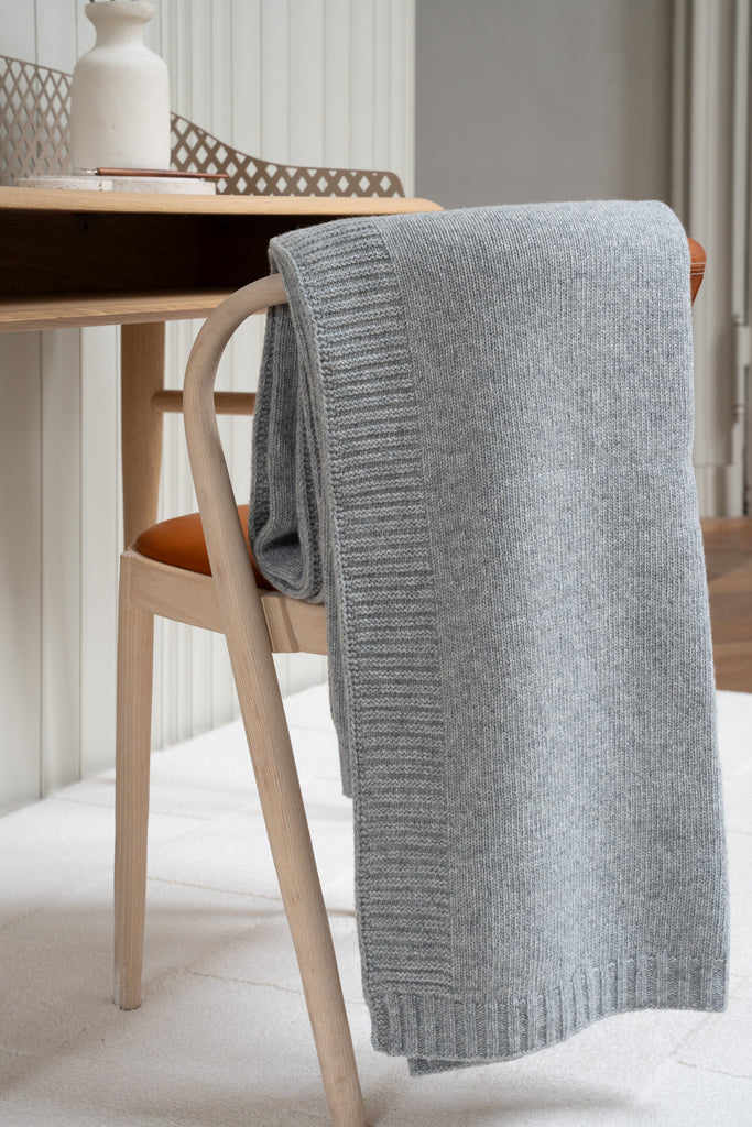 Gladgebreide grijze plaid van 100% merinowol, 130 x 170 cm, elegant over een stoel gehangen in Scandinavische interieur met een bureau op de achtergrond.