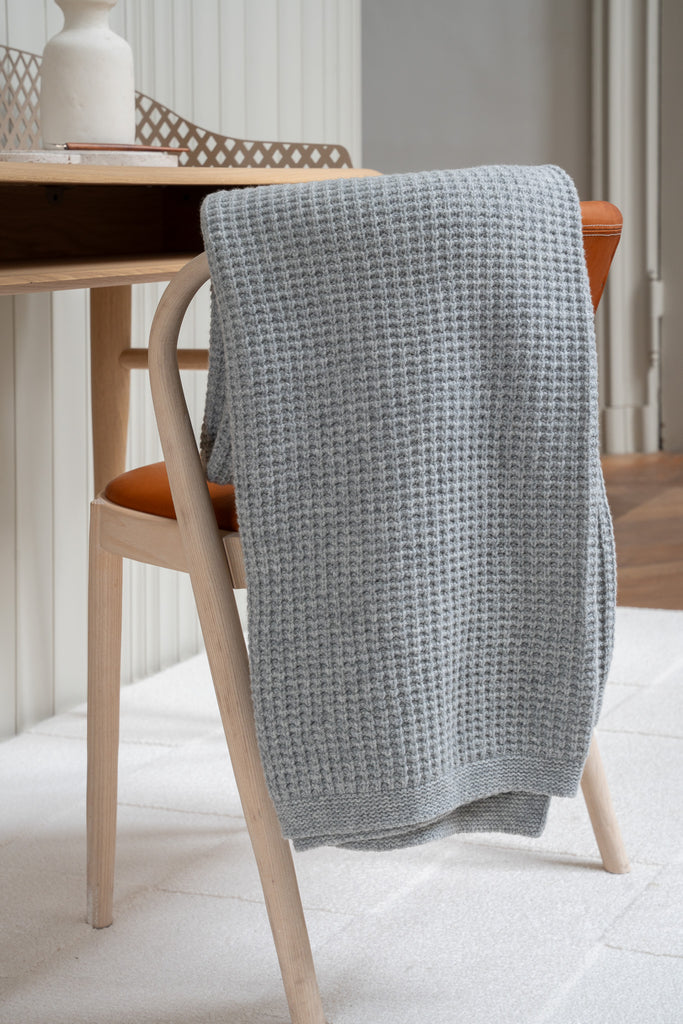 Grijze plaid van 100% merinowol met wafelpatroon, 130 x 170 cm, elegant over een stoel gehangen in Scandinavisch interieur met bureau op de achtergrond.