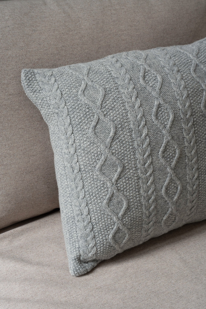 Sfeerbeeld van een grijs sierkussen 50 x 50 cm met Aran-kabelpatroon van 100% merinowol, subtiel geplaatst op een stijlvolle neutrale zetel.