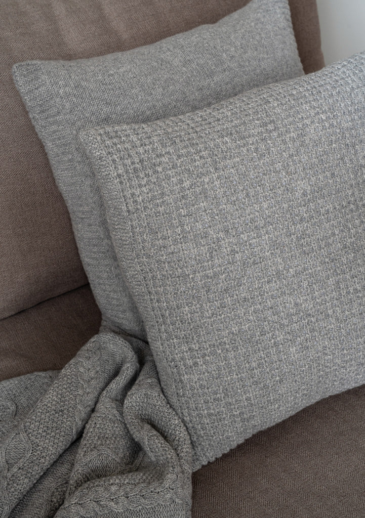 Grijze sierkussens van 100% merinowol met wafel- en glad breisel, subtiel geplaatst in een moderne zetel gemaakt van natuurlijke materialen.