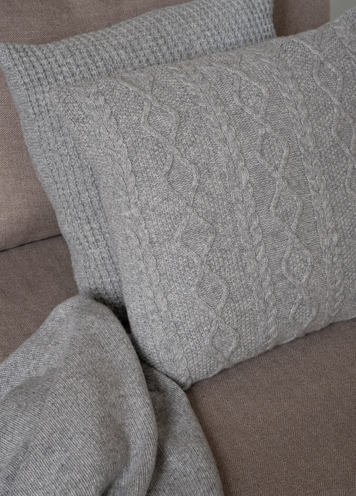 Sfeerbeeld van gestapelde grijze merinowollen sierkussens met wafel- en kabelpatronen (Aran), subtiel gestyled op een stijlvolle zetel.