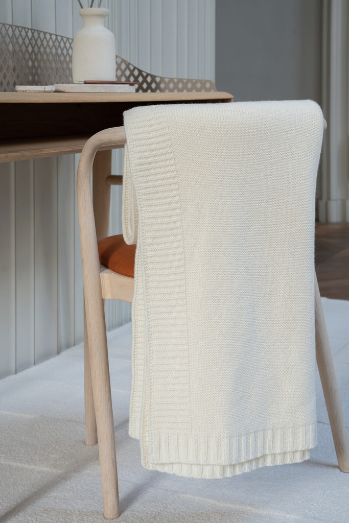 Gladgebreide witte plaid van 100% merinowol, 130 x 170 cm, elegant over een stoel gehangen in Scandinavische interieur met een bureau op de achtergrond.