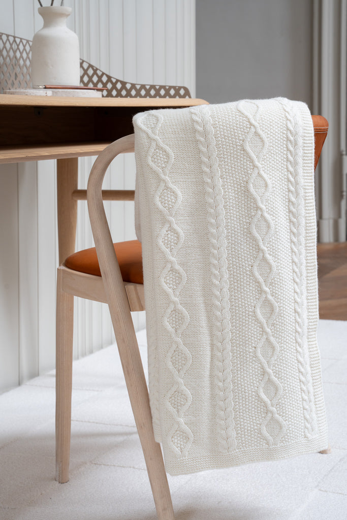Vlechtmotief witte plaid van 100% merinowol, 130 x 170 cm, elegant over een stoel gehangen in een Scandinavisch interieur met een bureau op de achtergrond.