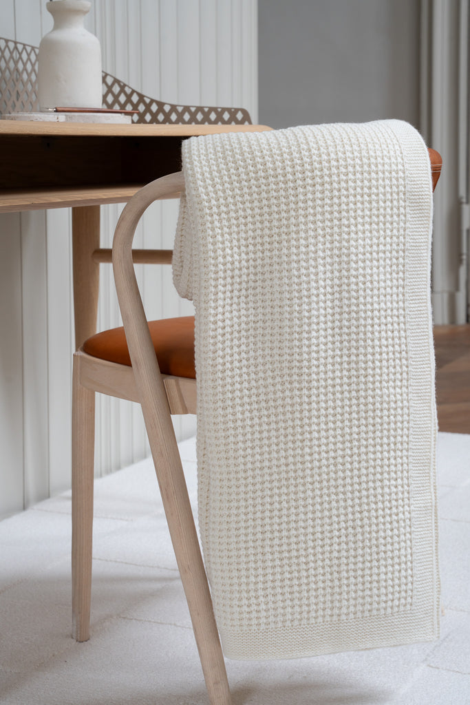 Witte plaid van 100% merinowol met wafelpatroon, 130 x 170 cm, elegant over een stoel gehangen in Scandinavisch interieur met bureau op de achtergrond.