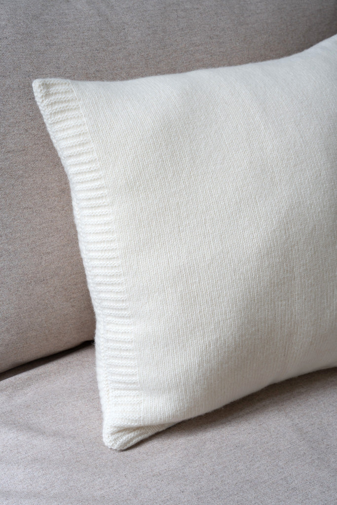Sfeerbeeld van een wit sierkussen met glad breisel van 100% merinowol,  50 x 50 cm, subtiel geplaatst op een neutrale zetel.